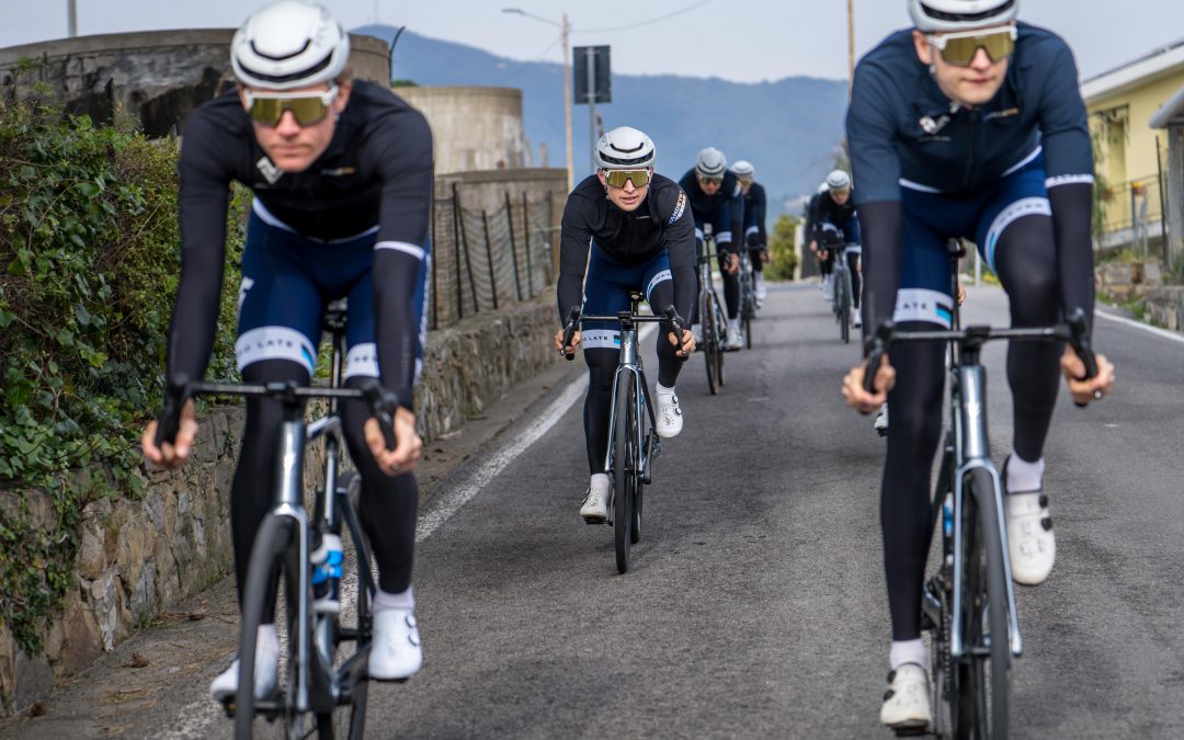 myvelo_rennrad_sanremo_web©invisions.at149 Eine Gruppe von Radfahrern in passender schwarzer und weißer Kleidung fährt auf einer asphaltierten Straße, mit Bergen und Gebäuden im Hintergrund. Die Radfahrer tragen Helme und Sonnenbrillen, und die Szene vermittelt ein Gefühl von Bewegung und Teamwork.