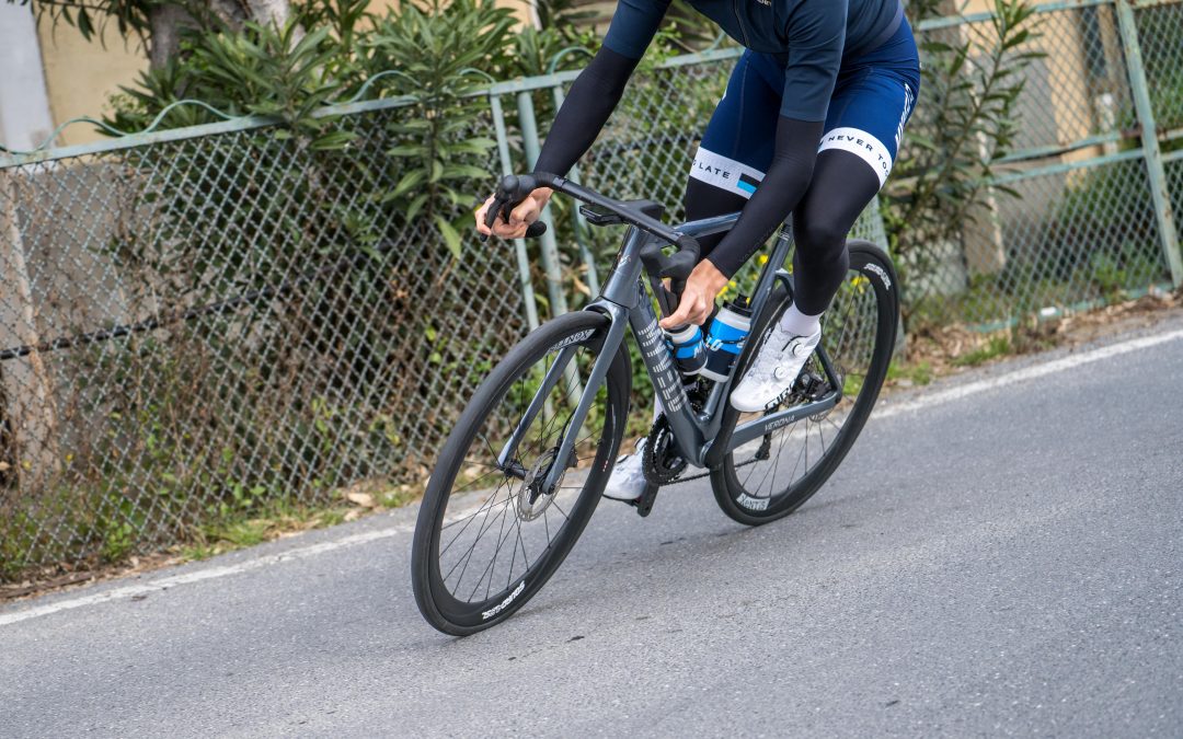 myvelo_rennrad_sanremo_web©invisions.at205 Ein Radfahrer in marineblauem Outfit und weißen Schuhen fährt auf einem schwarzen Rennrad auf einer gepflasterten Straße neben einem Maschendrahtzaun und grünen Sträuchern.