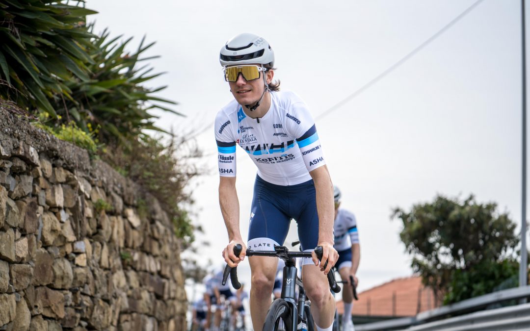 myvelo_rennrad_sanremo_web©invisions.at218 Ein Radfahrer in weiß-blauer Radfahrerkleidung, Helm und Sonnenbrille fährt auf einer Straße neben einer Steinmauer und Pflanzen, während im Hintergrund andere Radfahrer zu sehen sind.