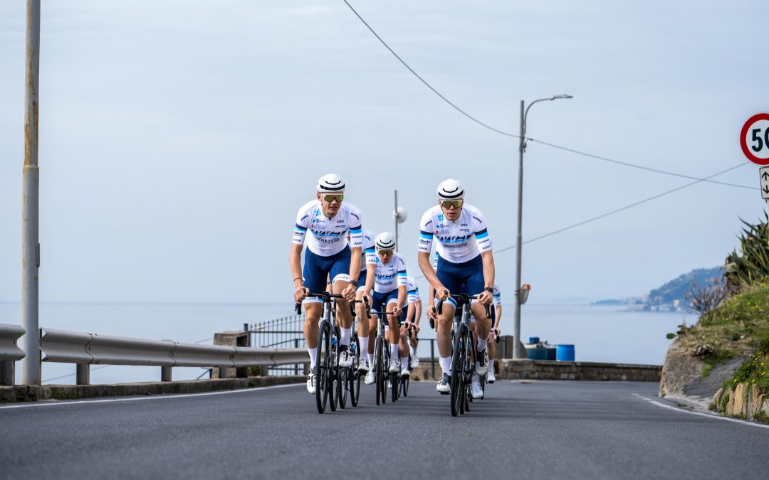 myvelo_rennrad_sanremo_web©invisions.at255 Eine Gruppe von fünf Radfahrern in passenden blau-weißen Uniformen fährt eng beieinander auf einer Küstenstraße, mit dem Meer und den Hügeln im Hintergrund. Ein Tempolimit-Schild und Geländer säumen die Straße.