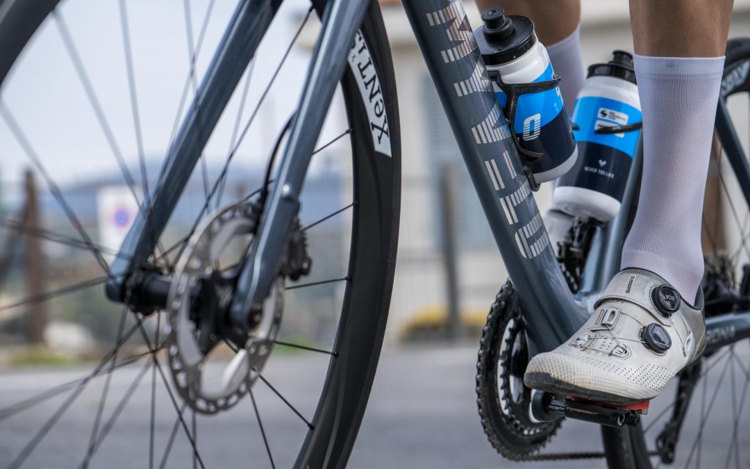 myvelo_rennrad_sanremo_web©invisions.at273 Nahaufnahme der Beine eines Radfahrers auf einem Rennrad, mit weißen Radschuhen, die in die Pedale eingeklinkt sind, zwei Wasserflaschen, die am Rahmen befestigt sind, und einem unscharfen Hintergrund im Freien.