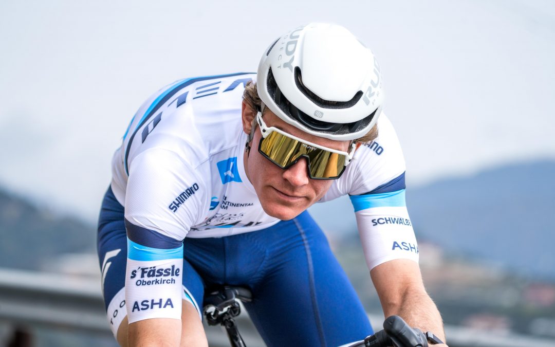 myvelo_rennrad_sanremo_web©invisions.at373 Ein Radfahrer, der einen weißen Helm, eine reflektierende Sonnenbrille und ein blau-weißes Rennoutfit trägt, fährt mit konzentriertem Blick im Freien. Der Hintergrund zeigt eine verschwommene Landschaft mit Hügeln und Himmel.