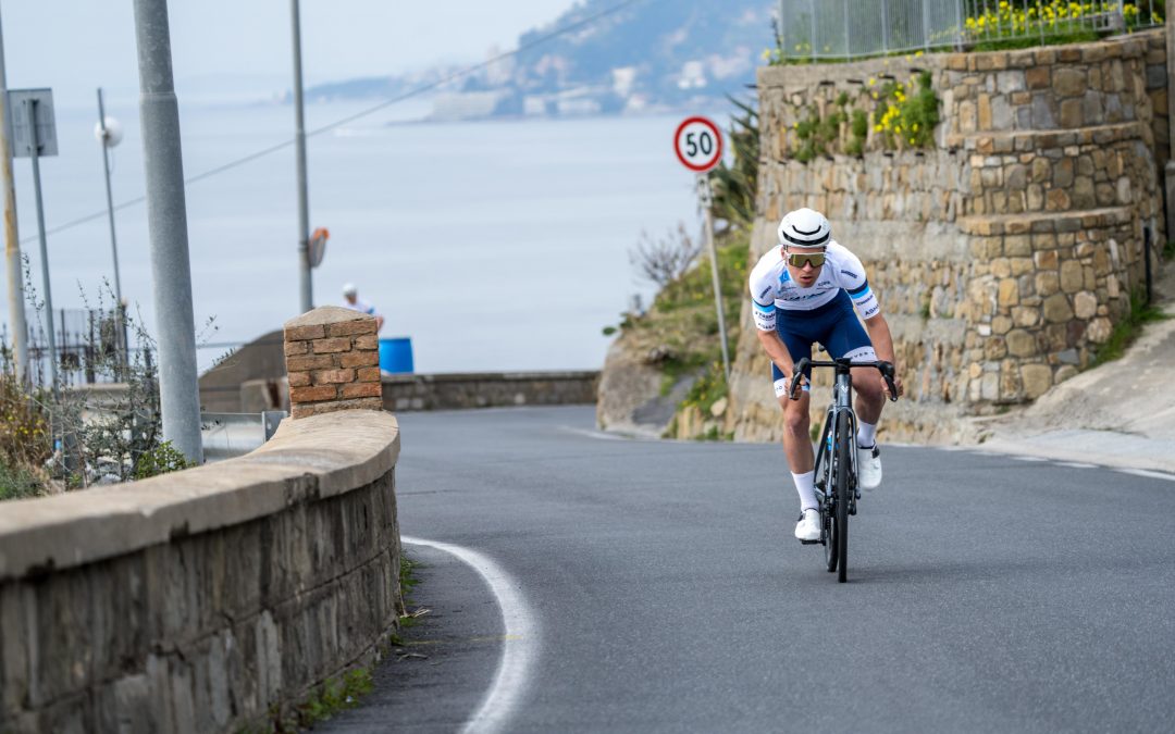 myvelo_rennrad_sanremo_web©invisions.at432 Ein Radfahrer mit gestreiftem Trikot und Helm fährt auf einer kurvenreichen Küstenstraße mit Steinmauern bergab, mit Blick auf das Meer und die fernen Hügel. Ein Schild mit einer Geschwindigkeitsbegrenzung von 50 km/h ist zu sehen.