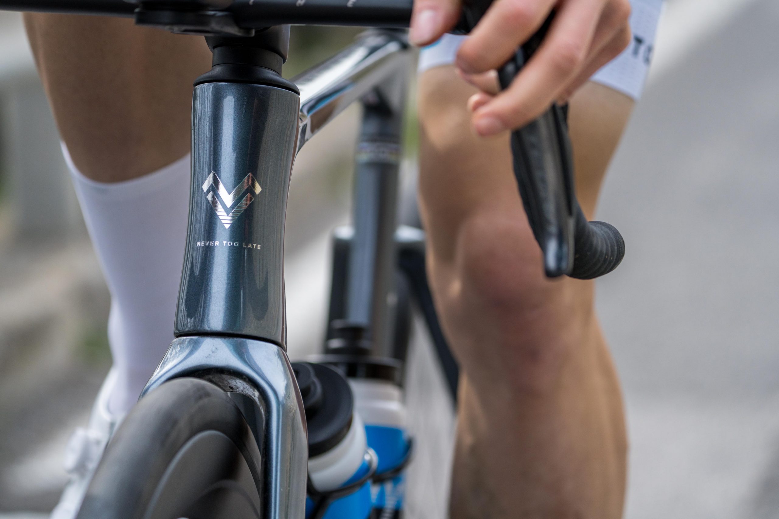 Nahaufnahme der Hand eines Radfahrers, der den Lenker eines Rennrads umgreift, wobei ein Teil des Fahrradrahmens, das Vorderrad, die Wasserflasche und das Knie des Radfahrers zu sehen sind. Auf dem Fahrradrahmen sind ein Logo und der Text "Never Too Late" zu sehen.