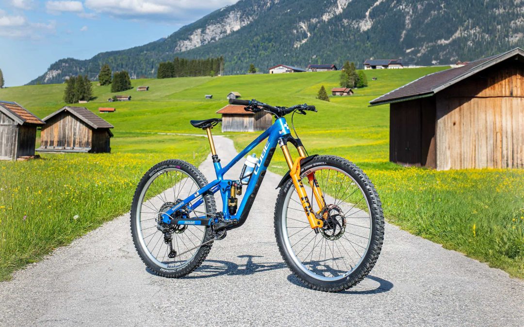 Ein blau-gelbes Mountainbike steht auf einem gepflasterten Weg auf einer grünen Wiese, umgeben von Holzhütten und im Hintergrund hohe Berge unter einem teilweise bewölkten Himmel.