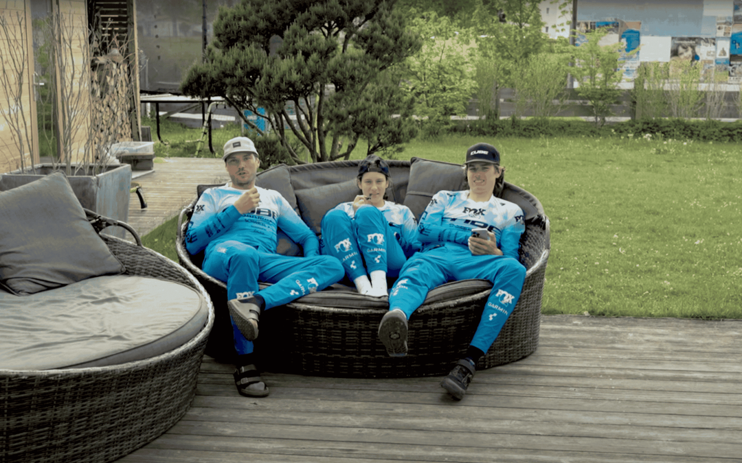 Drei Personen in passender blauer Sportkleidung entspannen sich auf einem runden Sofa im Freien auf einer Holzterrasse, mit Grünpflanzen und Bäumen im Hintergrund. Zwei von ihnen machen Friedenszeichen und einer hält ein Telefon in der Hand.