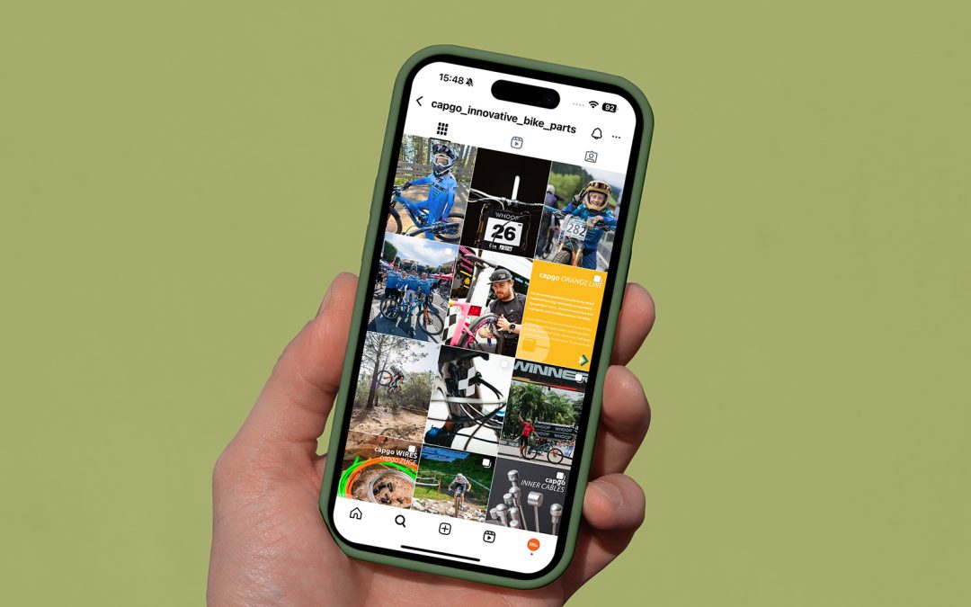 Eine Hand hält ein Smartphone, das ein Instagram-Profil mit einem Raster aus radsportbezogenen Beiträgen, darunter Radfahrer, Fahrräder und Werbegrafiken, vor einem schlichten olivgrünen Hintergrund anzeigt.