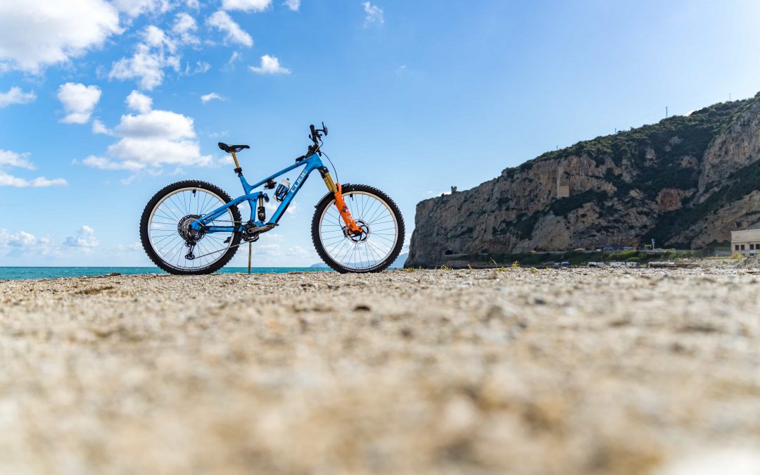 Ein blaues Mountainbike steht auf einer sandigen Fläche in der Nähe des Meeres, mit einem felsigen Abhang und einem teilweise bewölkten blauen Himmel im Hintergrund.