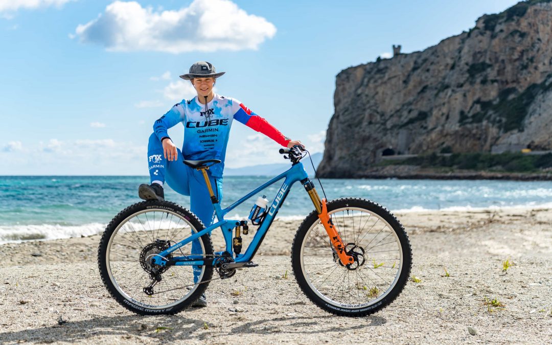 Eine Person in blauer Radfahrerkleidung posiert mit einem blauen Mountainbike an einem Strand, wobei das Meer und eine felsige Klippe im Hintergrund unter einem teilweise bewölkten Himmel zu sehen sind.