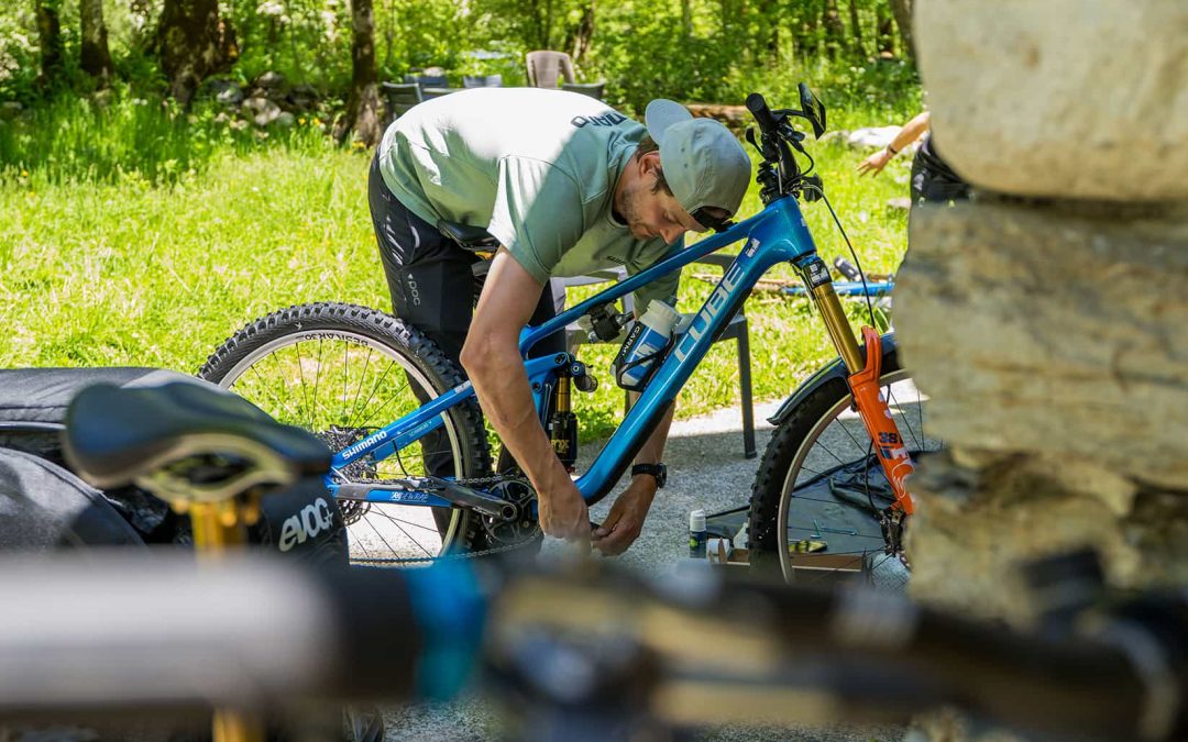 Eine Person mit umgekehrter Mütze bückt sich, um ein blaues Mountainbike im Freien an einem sonnigen Tag einzustellen, umgeben von grünem Gras und Bäumen, mit Fahrradausrüstung in der Nähe.