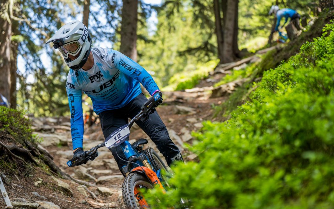 Ein Mountainbiker mit blau-weißem Trikot, Helm und Schutzbrille fährt einen felsigen Waldweg hinunter, während im Hintergrund zwischen hohen Bäumen und grünem Laub ein weiterer Radfahrer zu sehen ist.