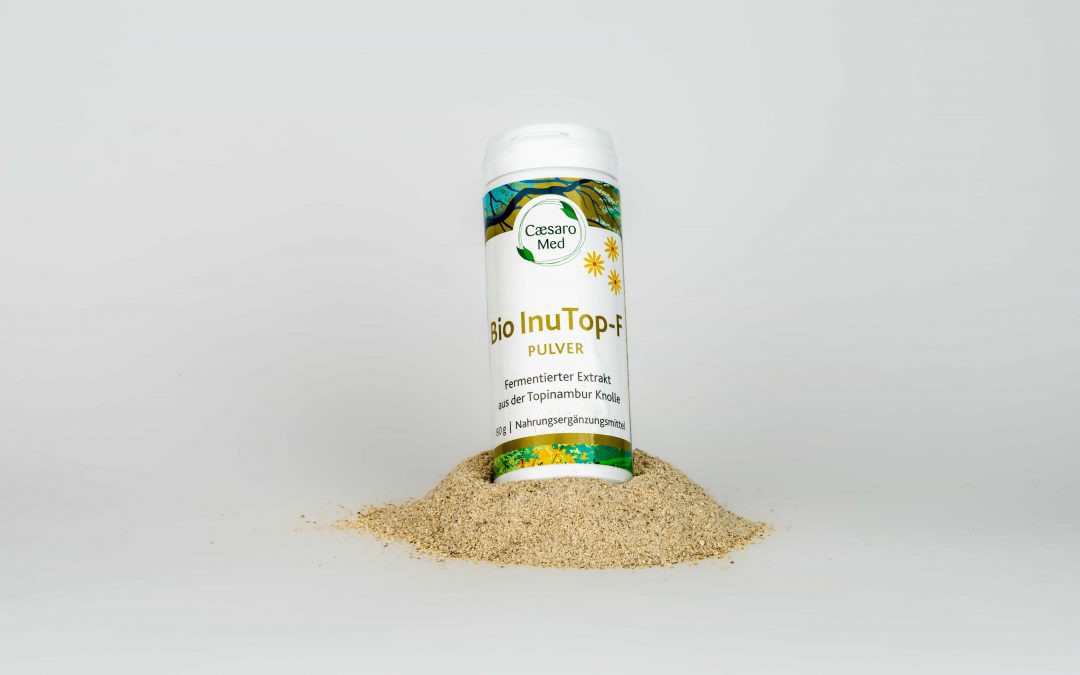 Ein weißer Behälter mit der Aufschrift Bio InuTop-F Pulver steht aufrecht in einem kleinen Hügel aus beigem Pulver vor einem schlichten weißen Hintergrund.