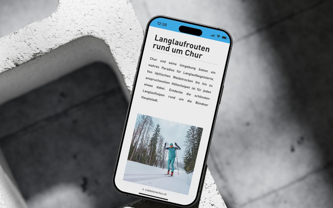 Ein Smartphone liegt auf einem weißen, strukturierten Block und zeigt eine deutsche Webseite mit dem Titel "Langlaufrouten rund um Chur" an, auf der ein Skifahrer in Winterausrüstung auf einer verschneiten Waldpiste zu sehen ist.