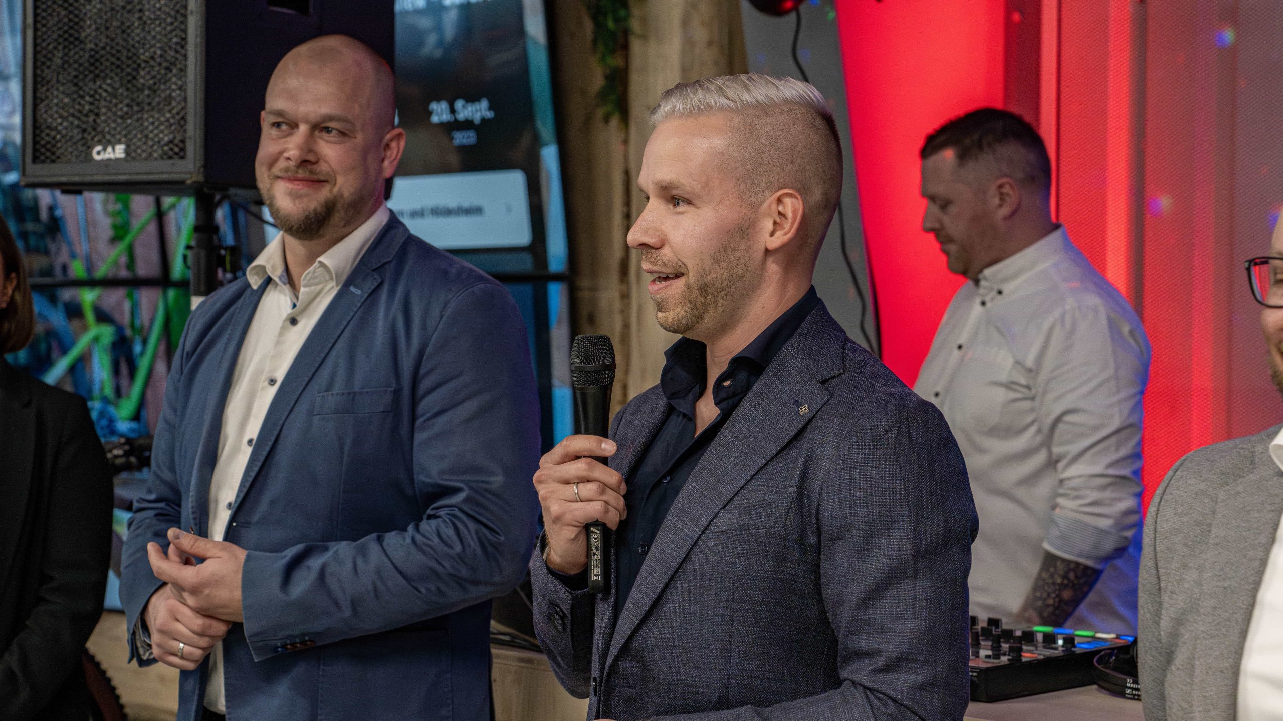 csgoslar_opening_36 Ein Mann in einem blauen Anzug spricht bei einer Indoor-Veranstaltung in ein Mikrofon, während ein anderer Mann in einem blauen Blazer und ein Mann in einem weißen Hemd in der Nähe stehen. Die rote und violette Beleuchtung schafft eine festliche Atmosphäre.