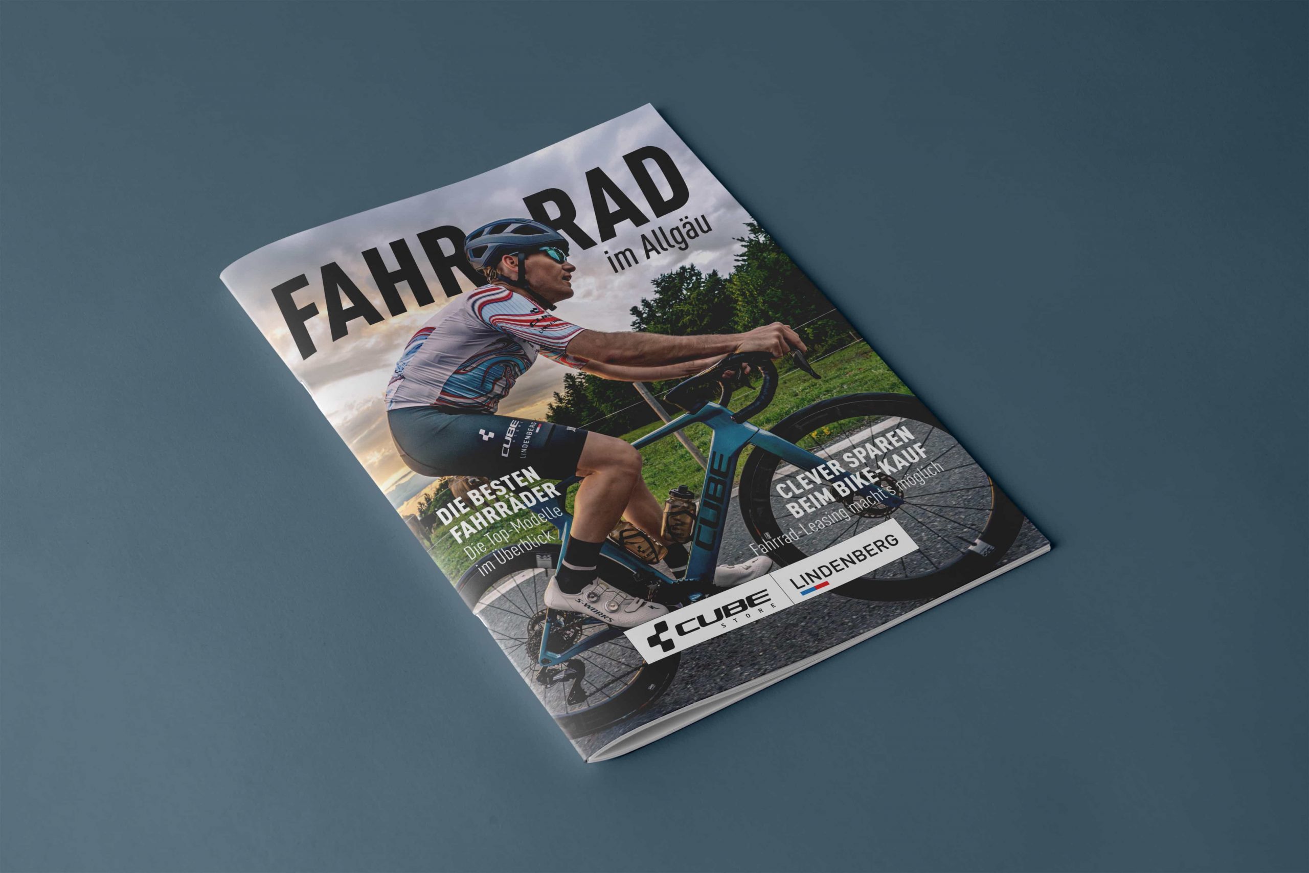 Eine Zeitschrift mit dem Titel FAHRRAD im Allgäu zeigt einen Radfahrer mit Helm und gestreiftem Trikot, der auf einer Straße fährt, mit einem malerischen, bewölkten Himmel und Bäumen im Hintergrund. Auf dem Umschlag befinden sich Werbetexte und Logos.