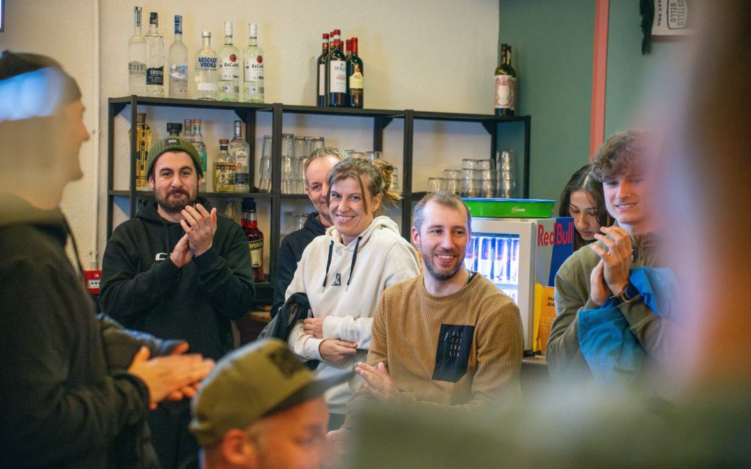 Eine Gruppe von Menschen in legerer Kleidung lächelt und applaudiert in einem Innenraum in der Nähe von Regalen mit Flaschen und einem Kühlschrank. Sie scheinen sich über ein Treffen oder eine Veranstaltung zu freuen.