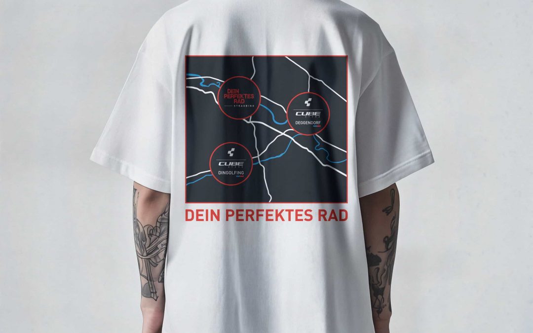 Eine Person trägt ein weißes T-Shirt mit einer Kartengrafik und drei markierten Orten auf dem Rücken. Unter der Landkarte ist in roter Farbe der Text DEIN PERFEKTES RAD aufgedruckt. Die Person hat tätowierte Arme.