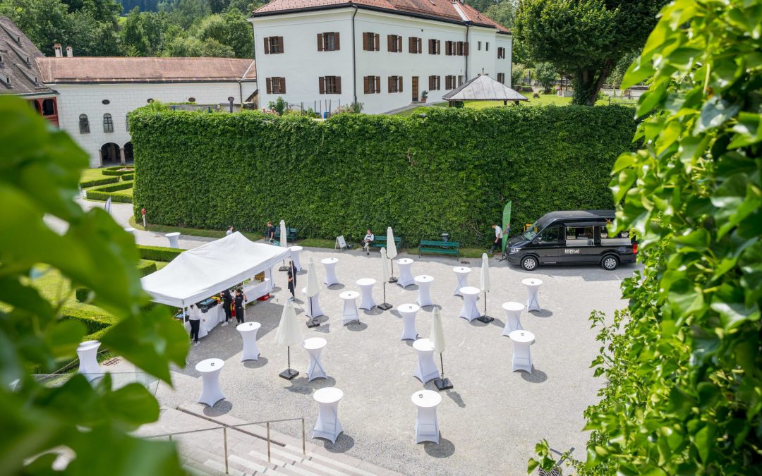 Ein Innenhof im Freien, der für eine Veranstaltung mit weißen Cocktailtischen, einem Zelt und einem schwarzen Fahrzeug hergerichtet wurde. Der Bereich ist von grünen Hecken und historischen Gebäuden umgeben, in denen sich einige Leute einrichten.