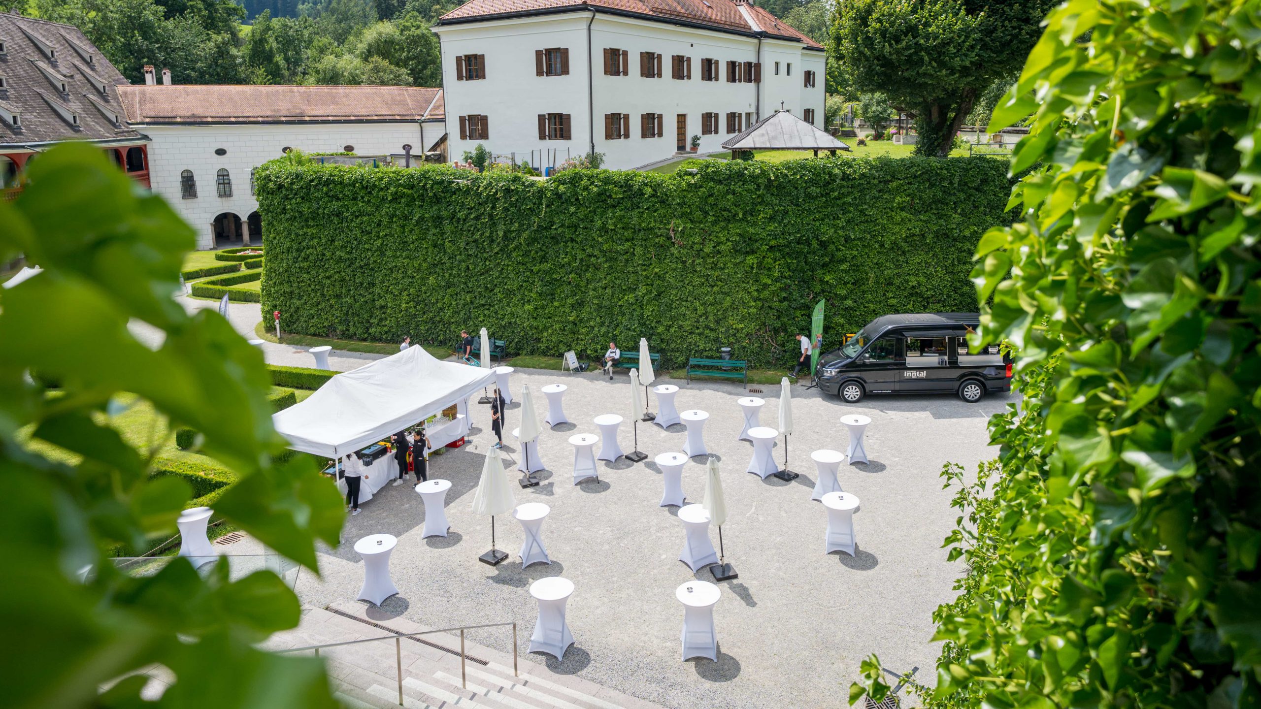 Ein Innenhof im Freien, der für eine Veranstaltung mit weißen Cocktailtischen, einem Zelt und einem schwarzen Fahrzeug hergerichtet wurde. Der Bereich ist von grünen Hecken und historischen Gebäuden umgeben, in denen sich einige Leute einrichten.