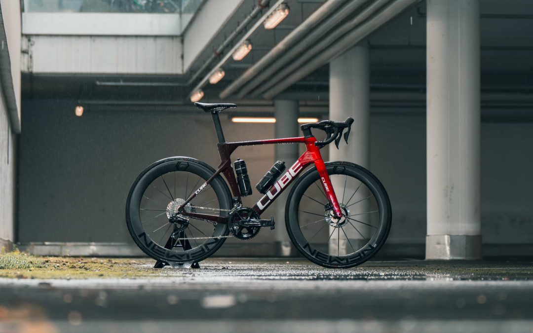 Ein rot-schwarzes Cube-Rennrad mit Tiefbettfelgen ist auf einer nassen Oberfläche in einem überdachten, städtischen Bereich mit Betonsäulen und Oberlicht abgestellt.
