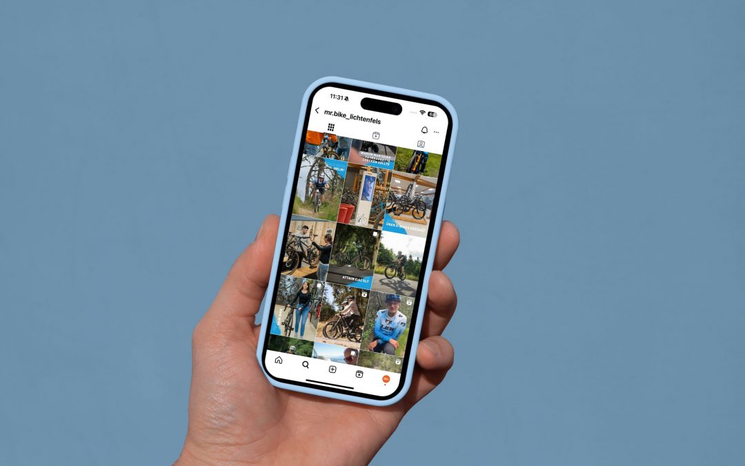 Eine Hand hält ein Smartphone, auf dem ein Instagram-Profil mit einer Reihe von Fotos zu sehen ist, die meist Fahrräder und eine Person im Freien vor einem einfarbig blauen Hintergrund zeigen.