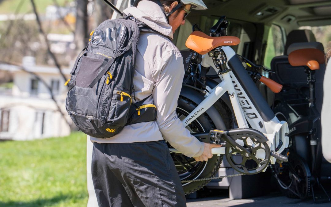 Eine Person, die einen Helm und einen Rucksack trägt, lädt an einem sonnigen Tag ein weißes Fahrrad mit orangefarbenen Akzenten auf die Ladefläche eines Fahrzeugs.