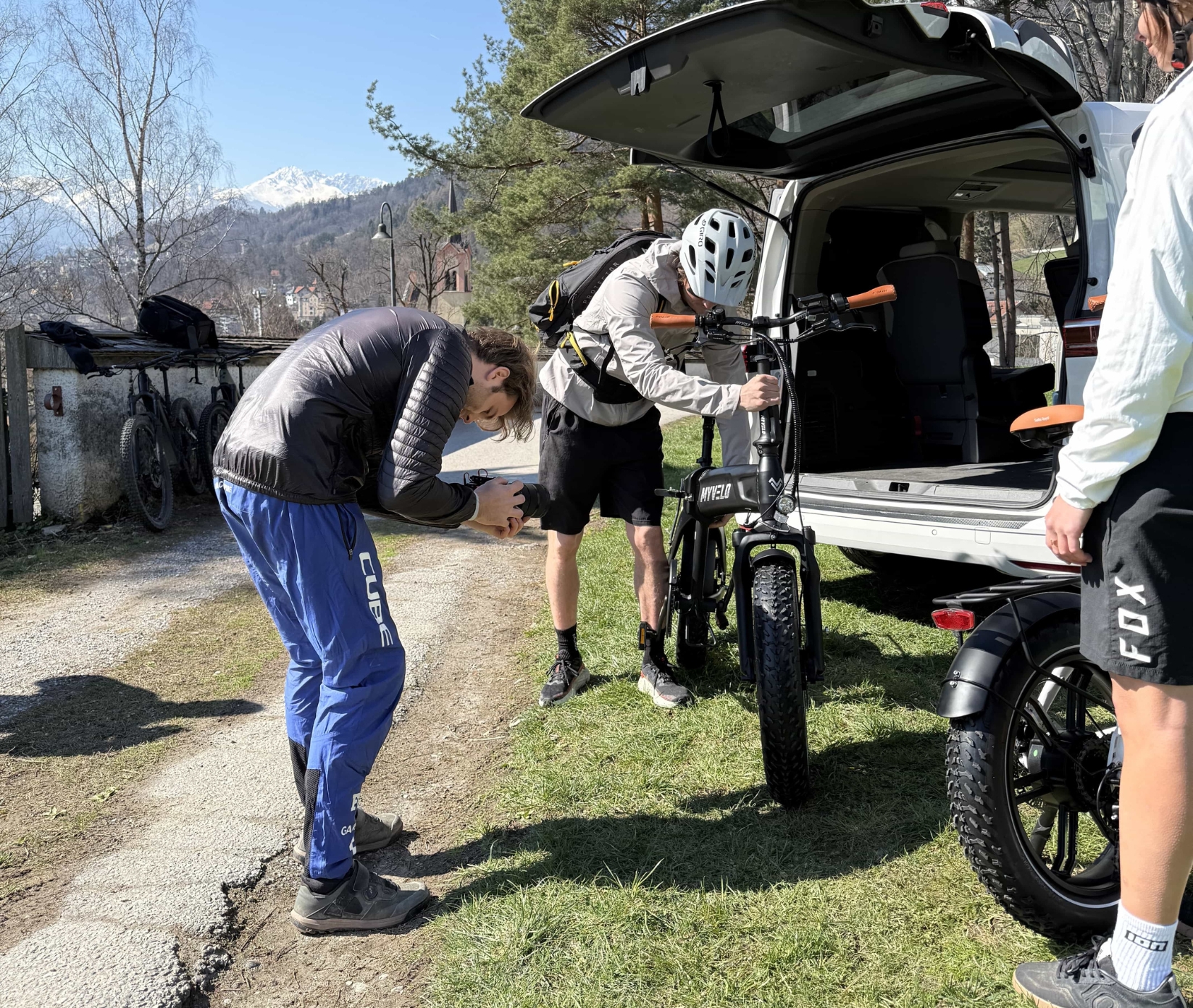 Zwei Personen laden an einem sonnigen Tag elektrische Mountainbikes aus dem Heck eines weißen Lieferwagens aus, während sich eine weitere Person in blauer Hose bückt, um eine Nahaufnahme zu machen. Im Hintergrund sind Bäume und Berge zu sehen.