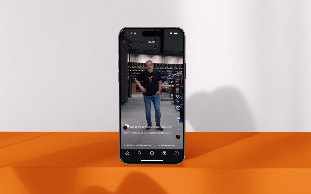 Ein Smartphone, das aufrecht auf einer orange-weißen Fläche steht, zeigt einen Social-Media-Post, der einen Mann in Freizeitkleidung zeigt, der in einem Haus steht, mit Regalen und Geräten im Hintergrund.