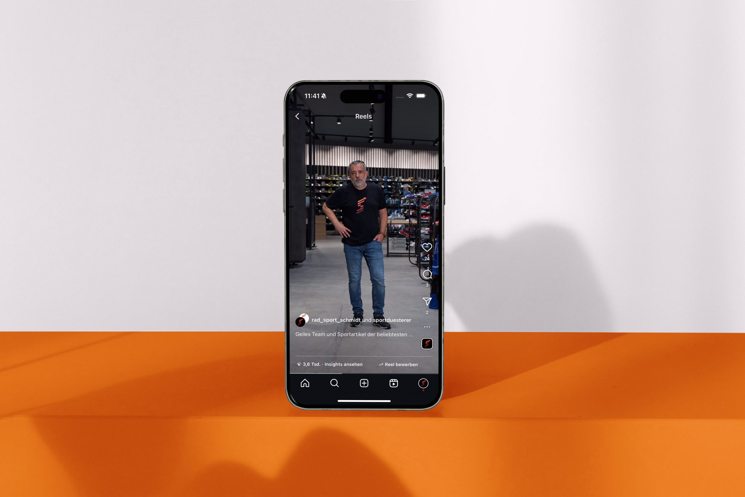 Ein Smartphone, das aufrecht auf einer orange-weißen Fläche steht, zeigt einen Social-Media-Post, der einen Mann in Freizeitkleidung zeigt, der in einem Haus steht, mit Regalen und Geräten im Hintergrund.