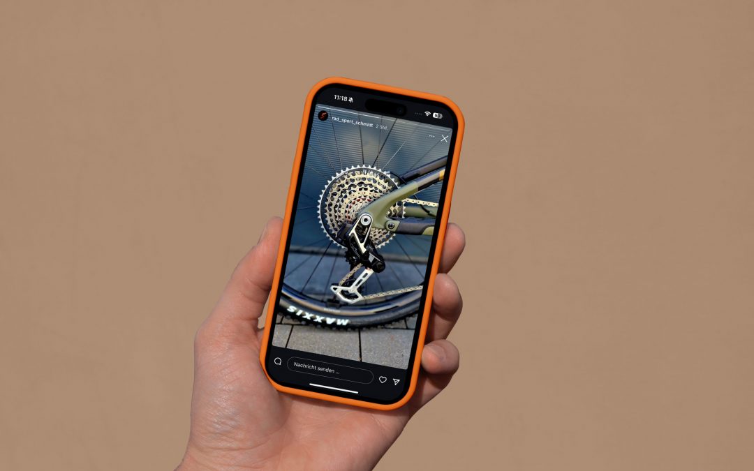 Eine Hand hält ein Smartphone mit einem orangefarbenen Gehäuse, auf dessen Bildschirm ein Foto der Gangschaltung eines Fahrrads vor einem schlichten beigen Hintergrund zu sehen ist.