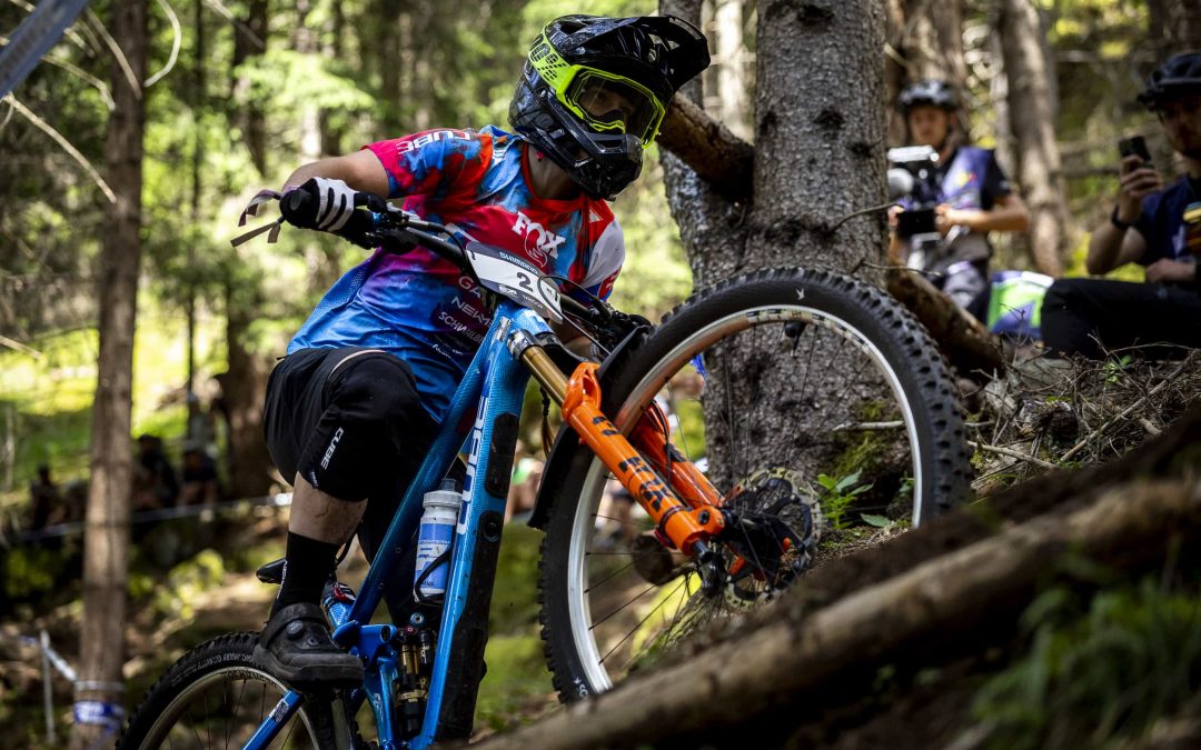 Ein Mountainbiker, der einen Helm und ein buntes Trikot trägt, fährt auf einem steilen, bewaldeten Weg, wobei er sich während eines Waldrennens konzentriert in eine Kurve legt. Im Hintergrund sehen Zuschauer zu.