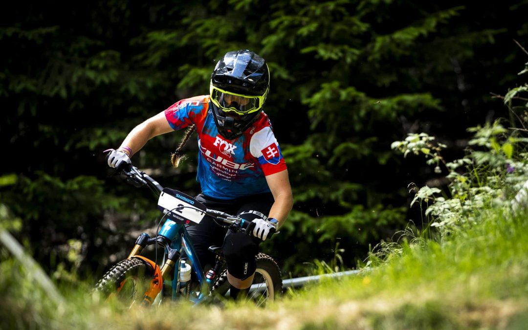 Ein Mountainbiker mit Helm, Schutzbrille und dem Trikot der slowakischen Mannschaft fährt bergab durch einen bewaldeten Weg, umgeben von Grün und Bäumen.