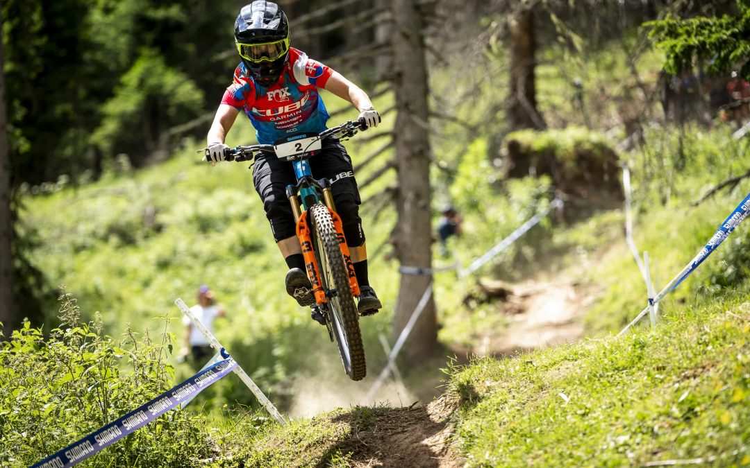 Ein Mountainbiker in voller Montur und mit Helm fährt über einen unbefestigten Waldweg, auf dem ein Band die Rennstrecke markiert und das Sonnenlicht durch die Bäume fällt.