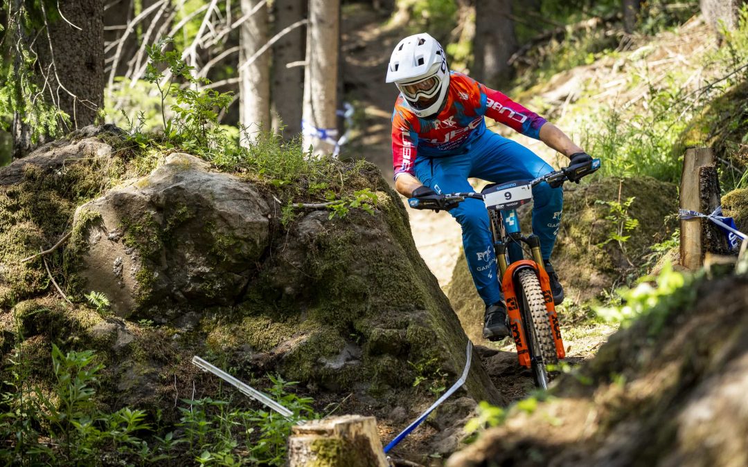Ein Mountainbiker in farbenfroher Kleidung und weißem Helm fährt bergab durch einen felsigen, bewaldeten Weg und manövriert zwischen Felsbrocken und Bäumen im hellen Sonnenlicht.