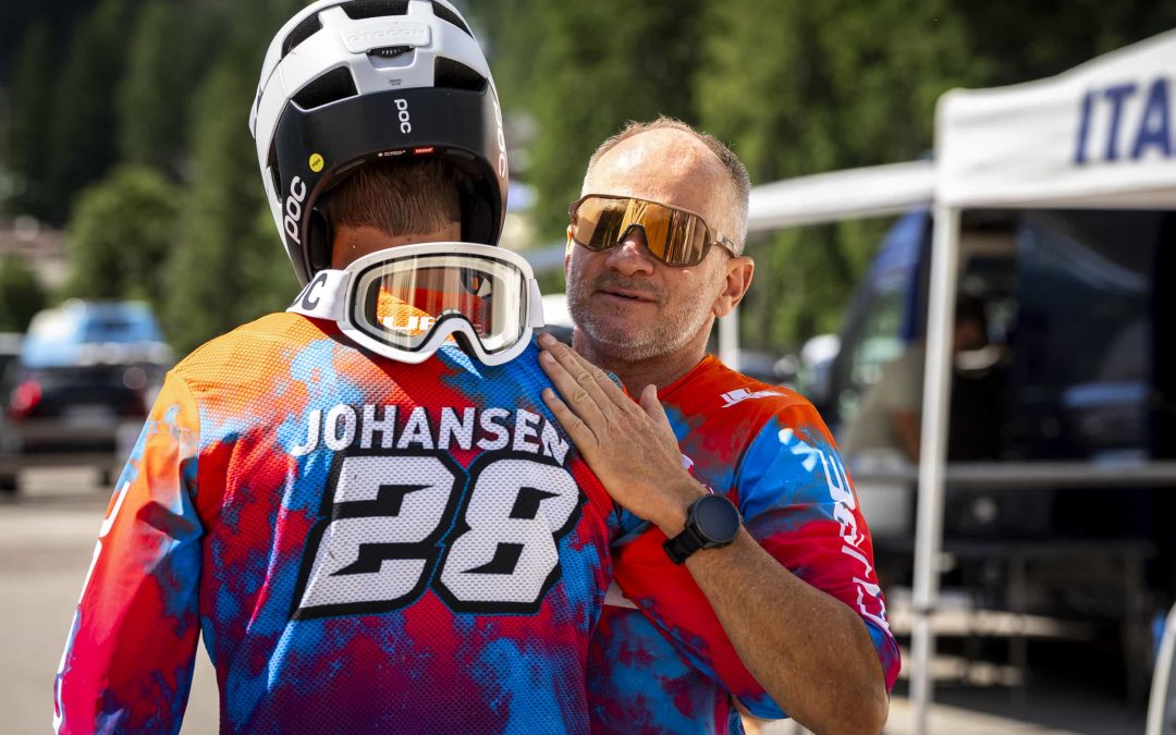 Ein Mann mit Sonnenbrille und buntem Trikot legt eine Hand auf die Schulter eines Radfahrers mit Helm und passendem Trikot mit der Aufschrift JOHANSEN 28, draußen in der Nähe von Bäumen und Zelten.