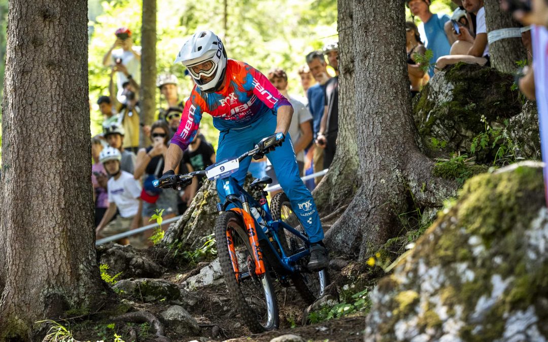 Ein Mountainbiker in einem rot-blau-weißen Outfit fährt auf einem felsigen Waldweg zwischen Bäumen hindurch, während im Hintergrund Zuschauer zusehen.