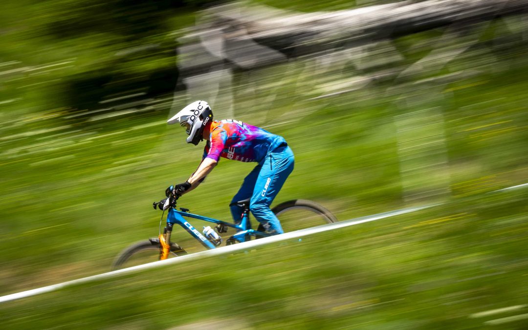 Ein Radfahrer mit weißem Helm und farbenfroher Ausrüstung fährt mit hoher Geschwindigkeit auf einem Mountainbike und erzeugt einen Bewegungsunschärfe-Effekt vor einem Hintergrund aus grünem Gras und Bäumen.