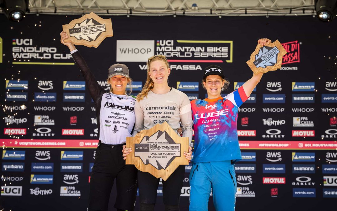 Drei Mountainbikerinnen stehen lächelnd auf einem Podium und halten hölzerne Trophäen über ihren Köpfen. Im Hintergrund sind die UCI Enduro World Series und verschiedene Sponsorenlogos zu sehen.