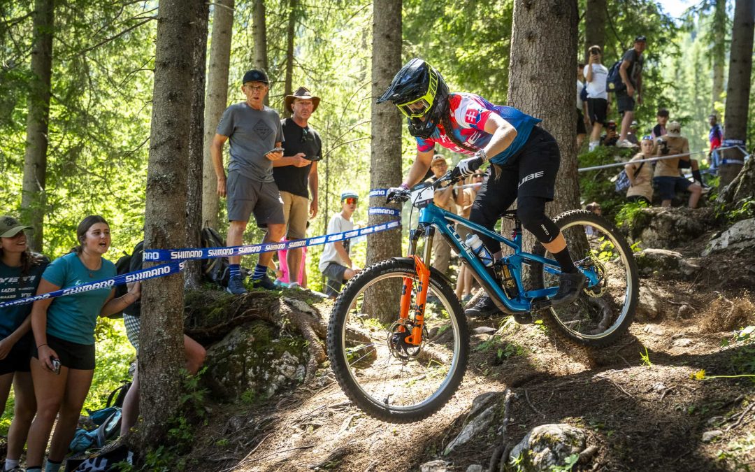 Ein Mountainbiker, der einen Helm und ein rot-blaues Trikot trägt, fährt während eines Rennens einen felsigen Waldweg hinunter, während Zuschauer hinter einer blauen Shimano-Schranke zwischen Bäumen zusehen.
