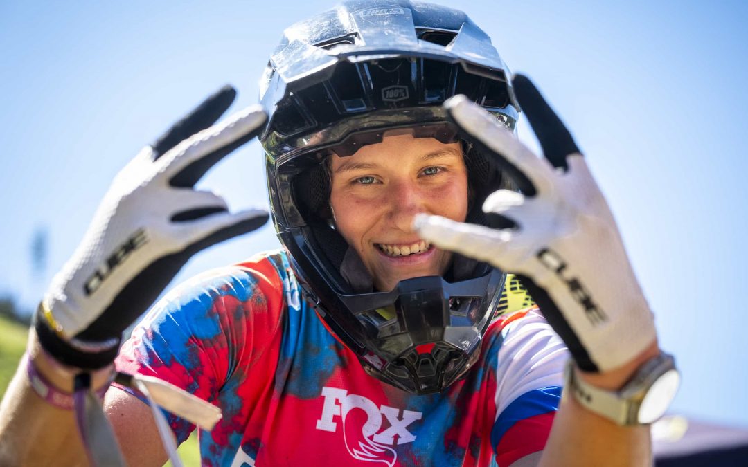 Eine Person, die ein buntes Trikot, Handschuhe und einen Vollvisierhelm trägt, lächelt in die Kamera und hält die behandschuhten Hände hoch, um mit ihren Fingern eine Form zu bilden. Der Himmel im Hintergrund ist klar und blau.