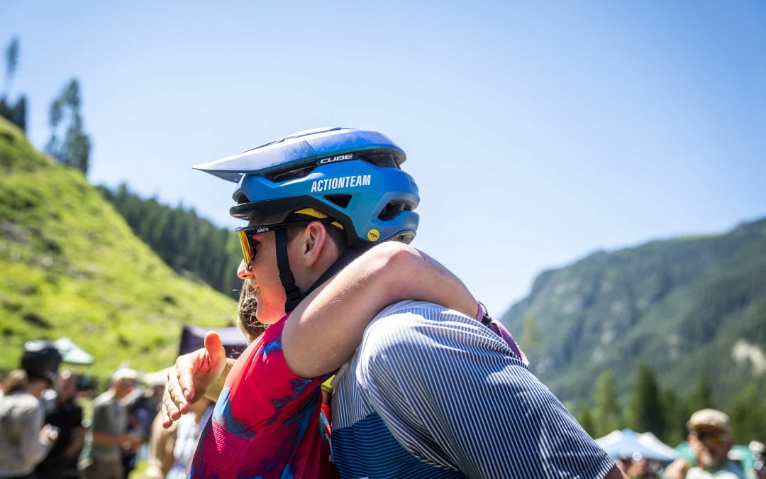 Zwei Radfahrer mit Helmen und bunten Trikots umarmen sich an einem sonnigen Tag im Freien, umgeben von grünen Hügeln, Bäumen und einem blauen Himmel. Menschen und Zelte sind im Hintergrund verschwommen.
