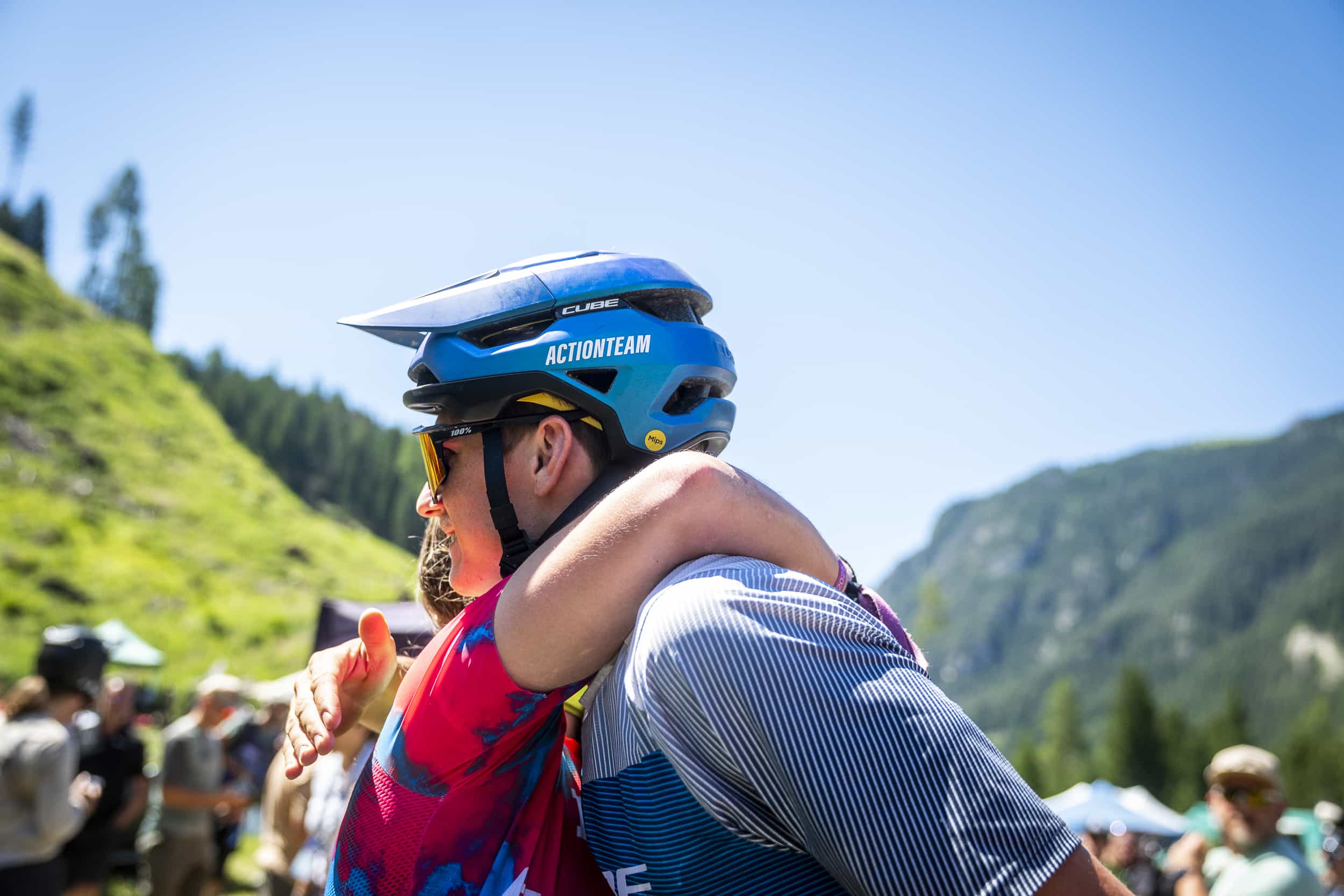 Zwei Radfahrer mit Helmen und bunten Trikots umarmen sich an einem sonnigen Tag im Freien, umgeben von grünen Hügeln, Bäumen und einem blauen Himmel. Menschen und Zelte sind im Hintergrund verschwommen.