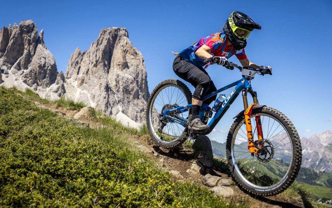 Ein Radfahrer mit Helm und farbenfroher Kleidung fährt mit seinem Mountainbike einen felsigen Weg hinunter, mit hohen, zerklüfteten Bergen und einem klaren blauen Himmel im Hintergrund.