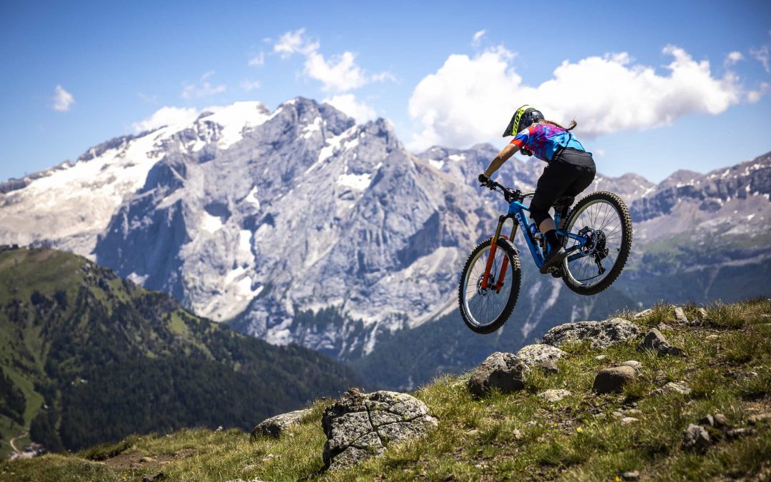 Ein Mountainbiker mit buntem Hemd und Helm springt von einem felsigen Hügel, mit schneebedeckten Bergen und einem blauen Himmel im Hintergrund.
