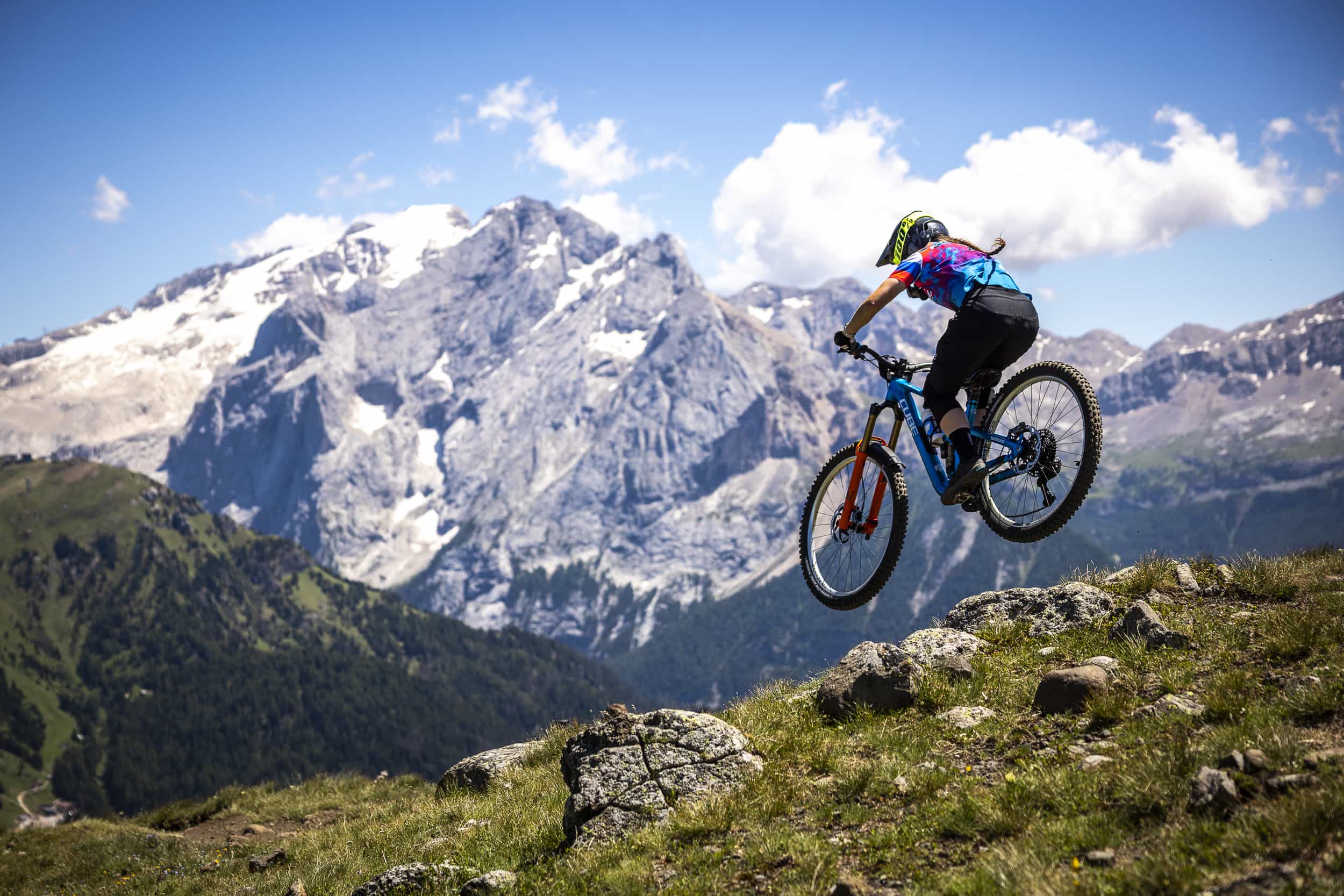 Ein Mountainbiker mit buntem Hemd und Helm springt von einem felsigen Hügel, mit schneebedeckten Bergen und einem blauen Himmel im Hintergrund.