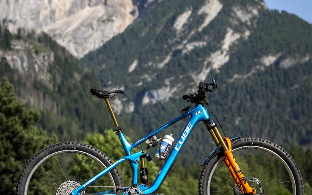 Ein hellblaues Cube-Mountainbike mit orangefarbener Vorderradaufhängung steht auf einer Wiese mit hohen, felsigen Bergen und grünem Wald im Hintergrund unter einem klaren blauen Himmel.
