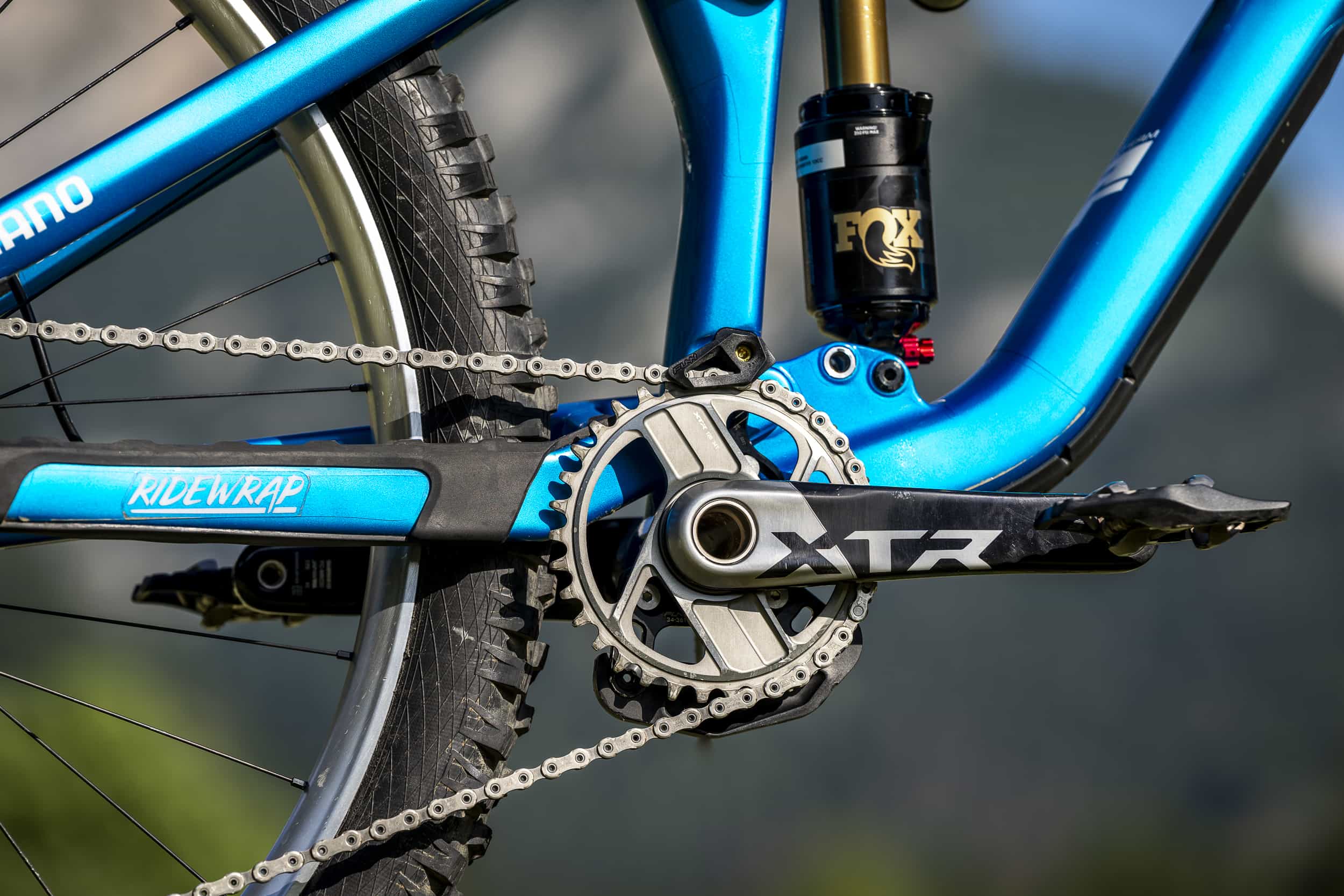 Nahaufnahme des Antriebsstrangs eines blauen Mountainbikes mit Shimano XTR-Kurbelgarnitur, Kettenblatt, Kette, Hinterradfederung und einem Teil des Hinterreifens vor einem unscharfen natürlichen Hintergrund.