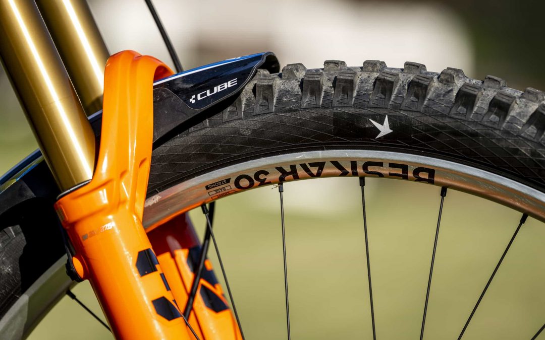Nahaufnahme eines Mountainbike-Vorderrads mit einem dicken schwarzen Reifen, einer orangefarbenen Federgabel und einem Teil der Felge mit der Aufschrift BESKAR 30. Der Hintergrund ist unscharf.