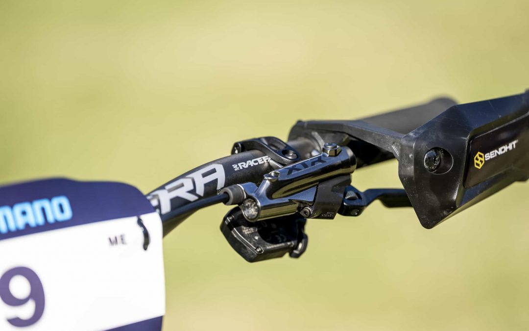 Nahaufnahme eines schwarzen Mountainbike-Lenkers mit einem SRAM-Bremshebel, einem Nummernschild mit der Zahl 9 und einem Handschutz mit dem Sendhit-Logo vor einem verschwommenen grünen Hintergrund.