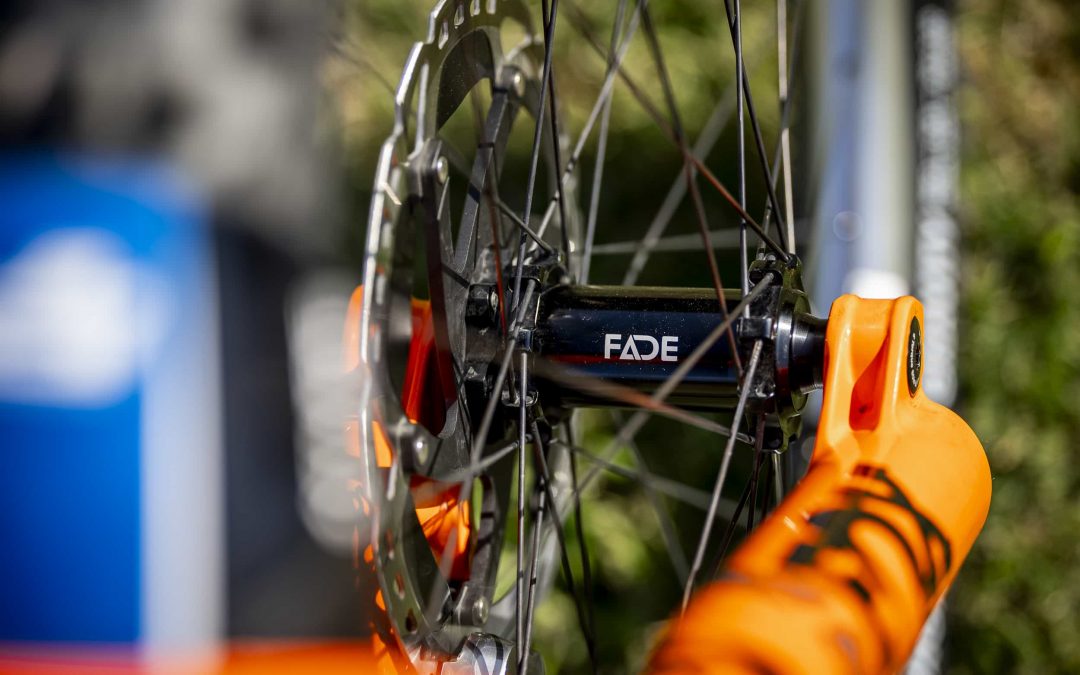 Nahaufnahme eines Fahrradrads mit Schwerpunkt auf dem Rotor der Scheibenbremse, der schwarzen Nabe mit der Aufschrift FADE und der orangefarbenen Gabel, mit unscharfem grünem Gras im Hintergrund.